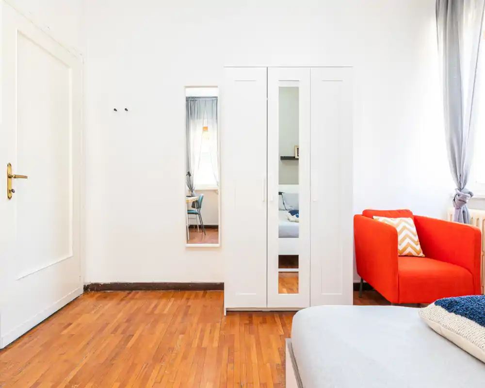 Bright, modern bedroom with a visible bed corner, white wardrobe with mirrored door, and a vivid orange armchair adding a pop of color.