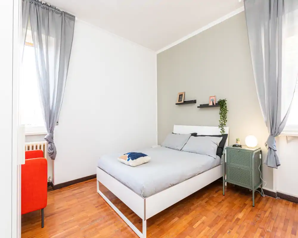 Bright, minimalist bedroom with a centrally placed double bed, soft grey linens, bedside cabinet and natural daylight from two windows.