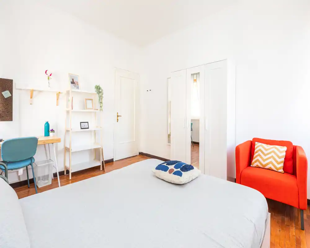 Bright, well-staged bedroom with a large bed in the foreground, a cozy red armchair, wardrobe with mirror and a small desk area — ideal for single or couple stays.