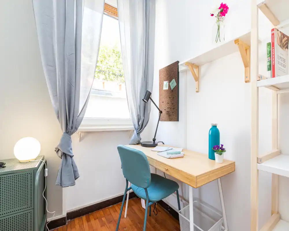 Bright, tidy bedroom corner with a wooden desk by the window, a turquoise chair, lamp and small decorative items — ideal for study or remote work.