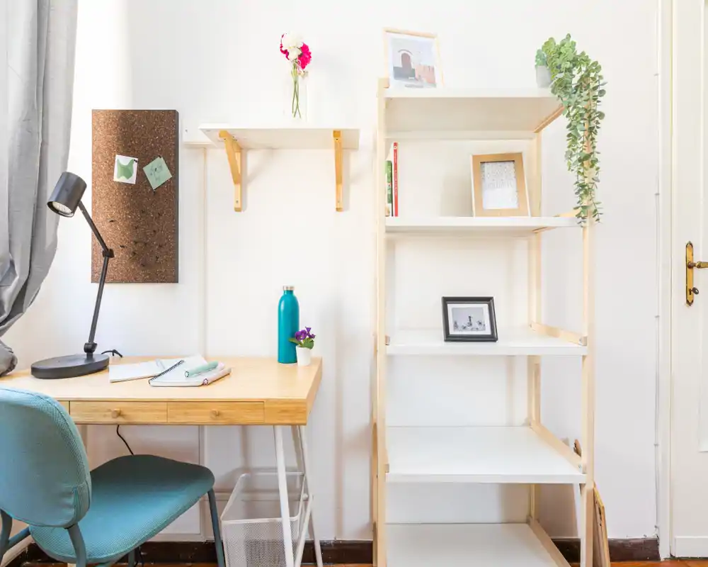 Bright, minimal workspace corner with a wooden desk, teal chair and open shelving; tidy styling with small plants and framed photos makes it welcoming.