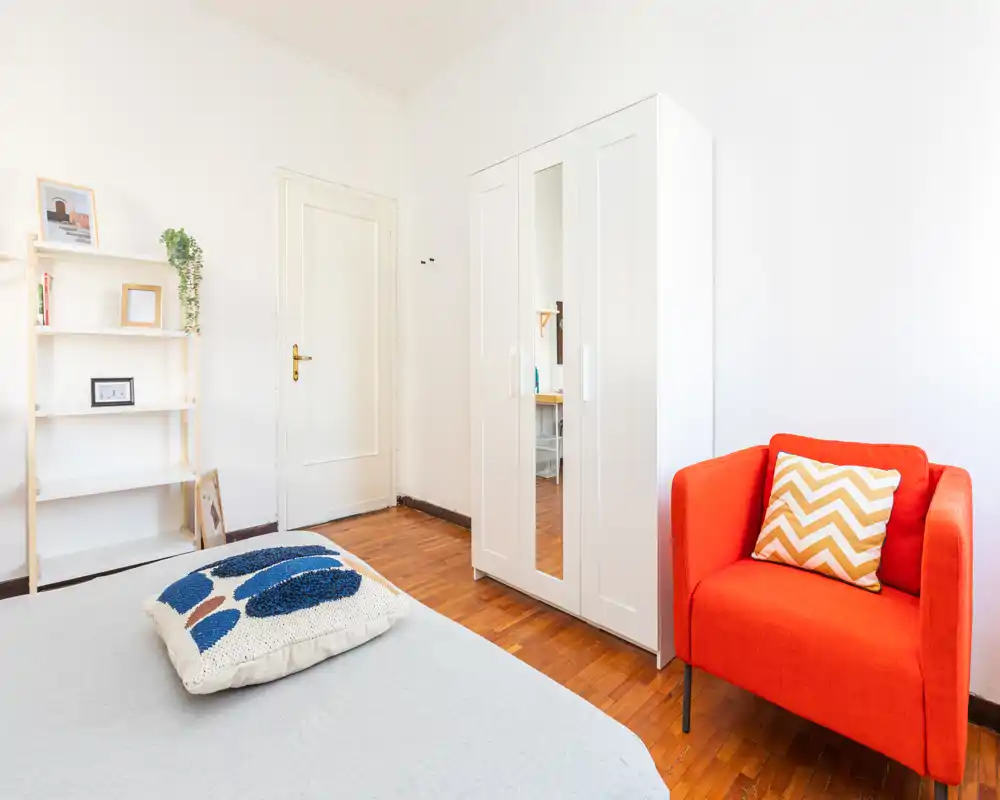 Bright, minimal bedroom with a visible bed, white wardrobe and a vibrant orange armchair adding a modern accent.