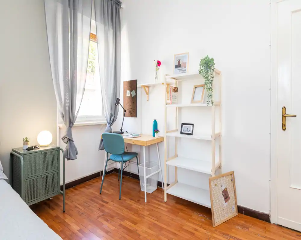 Bright, cozy bedroom corner with a small wooden desk and shelving beside a curtained window. Natural light, potted plant accents and warm wood flooring create a welcoming workspace within the sleeping area.