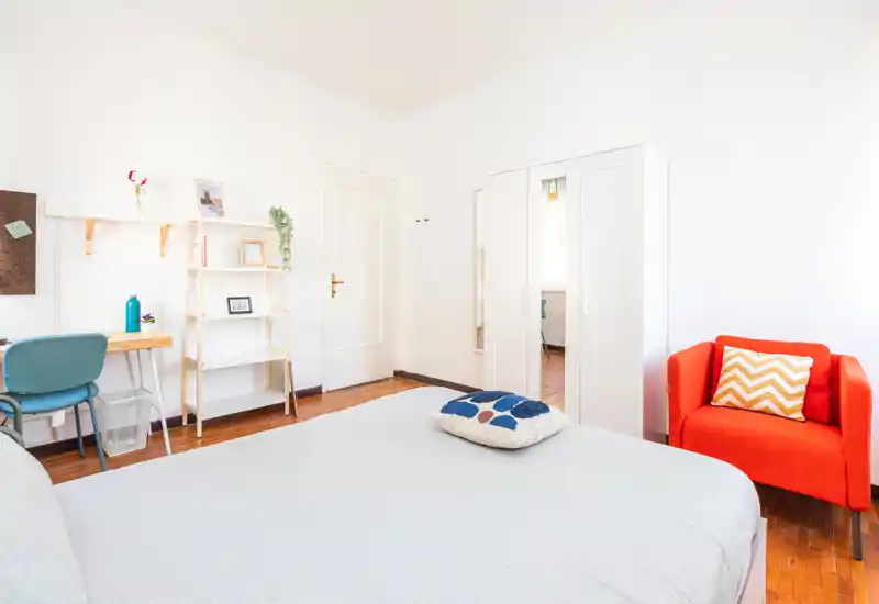 Bright, well-staged bedroom with a large bed in the foreground, a cozy red armchair, wardrobe with mirror and a small desk area — ideal for single or couple stays.
