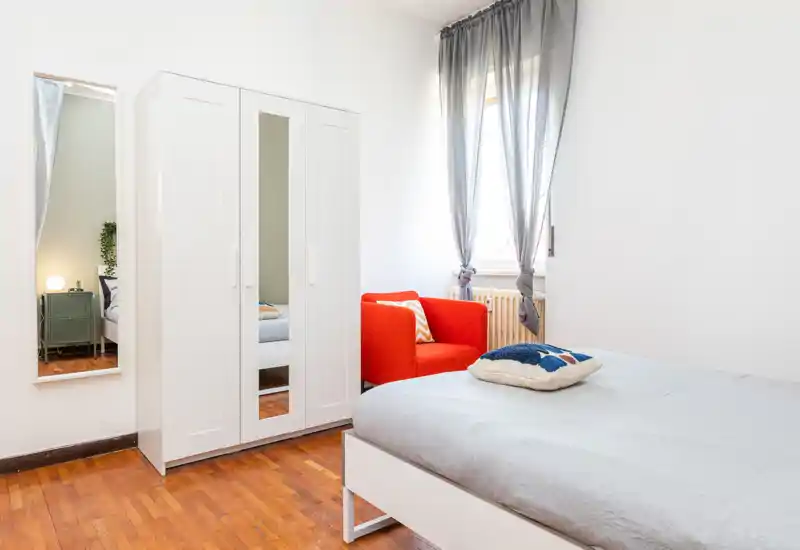 Bright, modern bedroom with a neatly made bed, a white wardrobe with mirrored door and a vivid orange armchair by the window — clean lines and warm wooden flooring give a welcoming feel.