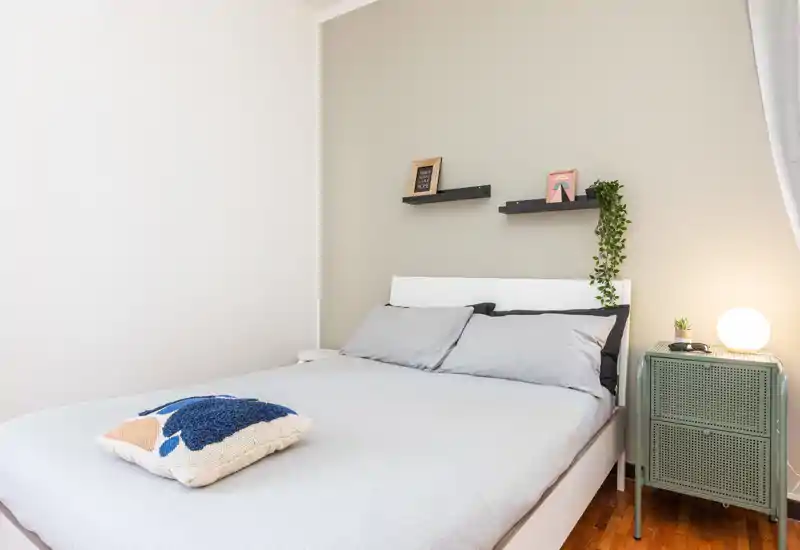 Bright, modern bedroom with a neatly made double bed, soft gray linens, decorative pillow and a bedside cabinet with lamp—minimal, cozy styling ideal for rental listings.
