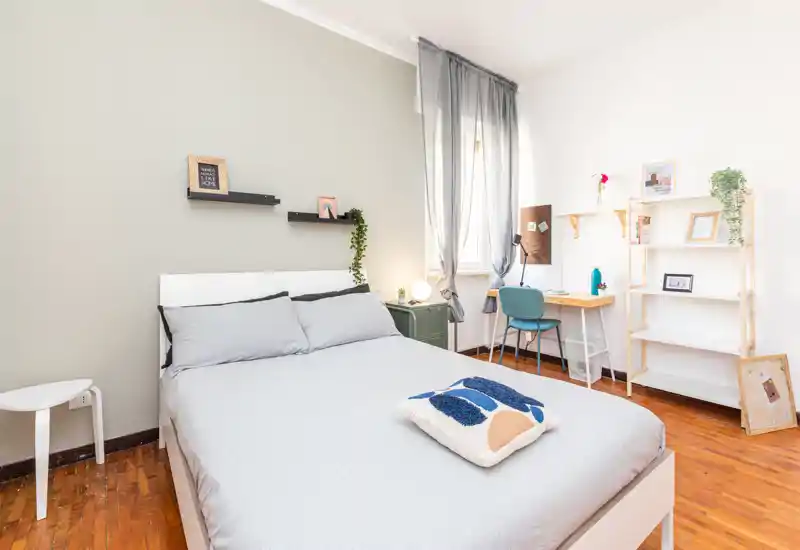 Bright, modern bedroom with a full double bed, soft gray linens, a small bedside table and a study corner by the window. Minimal, cozy decor with plants and open shelving creates a calm, welcoming atmosphere ideal for rental photos.