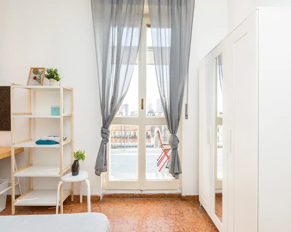 Chambre lumineuse et minimaliste avec grande fenêtre et voilages donnant sur un petit balcon; étagère simple, armoire avec miroir et coin lit confortable.
