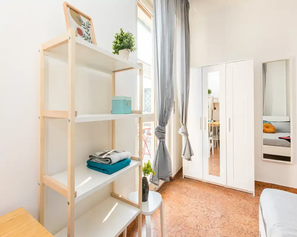 Coin de chambre lumineux et sobrement meublé avec une armoire, une étagère et une partie du lit visible. Lumière naturelle et tons neutres pour un espace propre et attractif pour les annonces locatives.