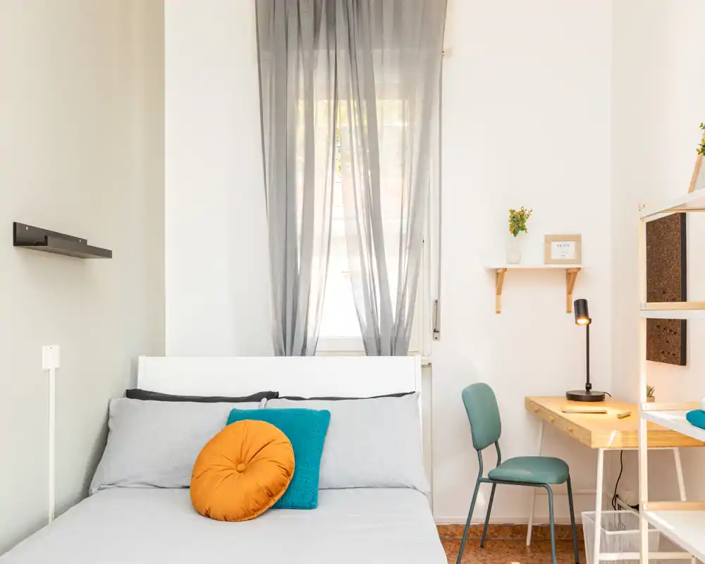 Chambre lumineuse et bien arrangée avec un lit entièrement visible, coussins colorés et un petit bureau près de la fenêtre — idéale pour les locations courte durée au style moderne et minimaliste.