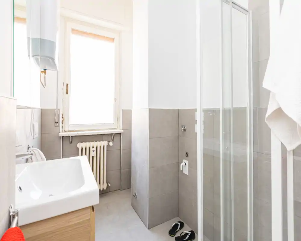 Bright, modern bathroom with a glass shower enclosure, wall-mounted radiator, sink with wooden cabinet and natural light from a window.