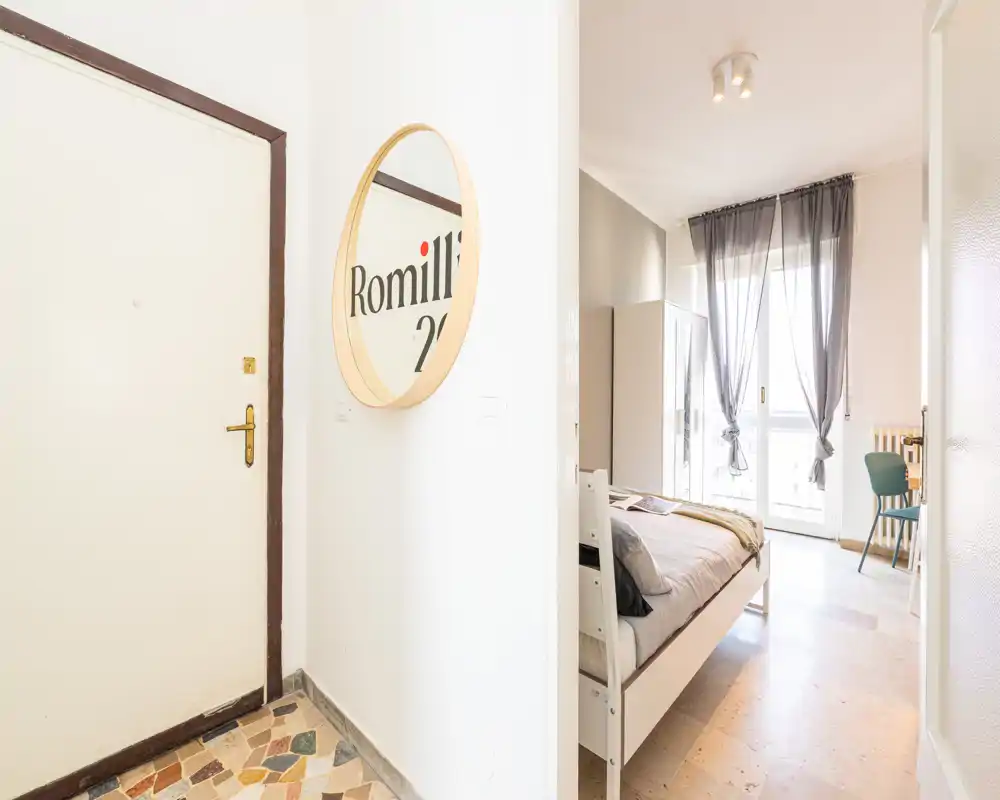 Bright, airy bedroom seen through an entrance: a neat single bed, wardrobe and sheer curtains opening to a balcony provide a fresh, minimalist feel.