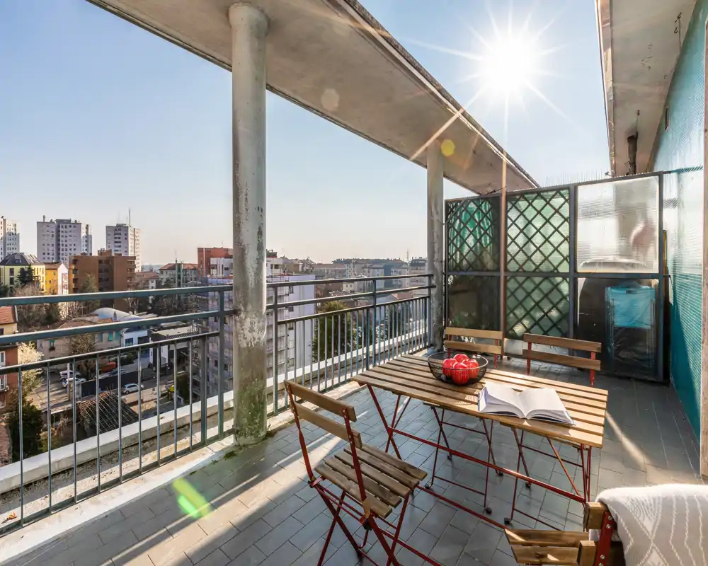 Sunny furnished balcony with a small wooden table and folding chairs, open city views and protective railing — ideal for morning coffee.