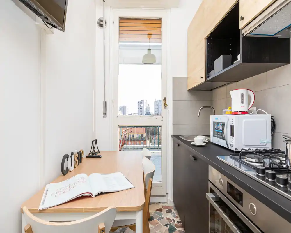 Compact modern kitchen with a wooden dining table, gas hob, microwave and natural light from a balcony door — ideal for a city apartment.