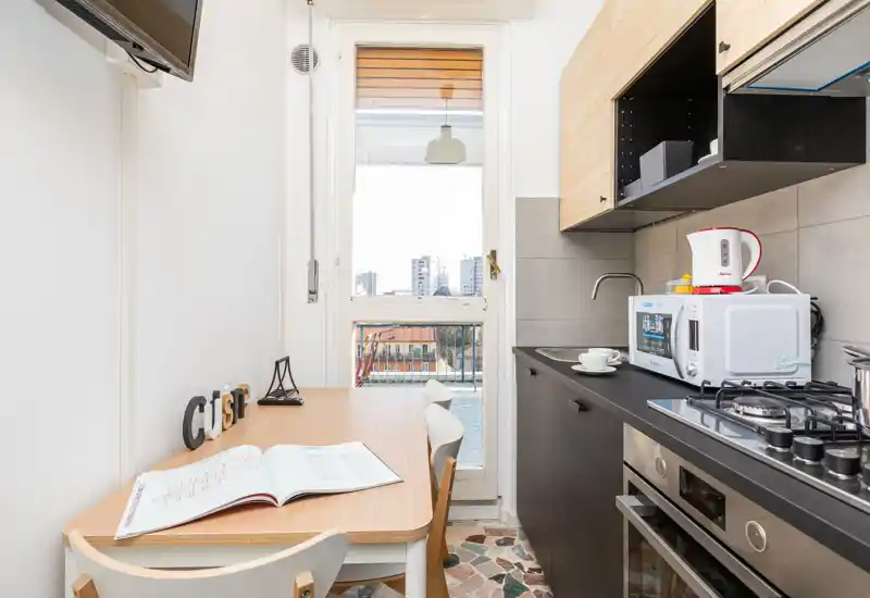 Compact modern kitchen with a wooden dining table, gas hob, microwave and natural light from a balcony door — ideal for a city apartment.