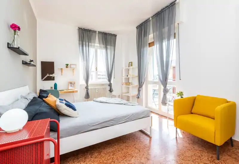 Camera da letto luminosa e moderna con ampio letto bianco, biancheria morbida, cuscini decorativi e una poltrona gialla davanti al balcone soleggiato.