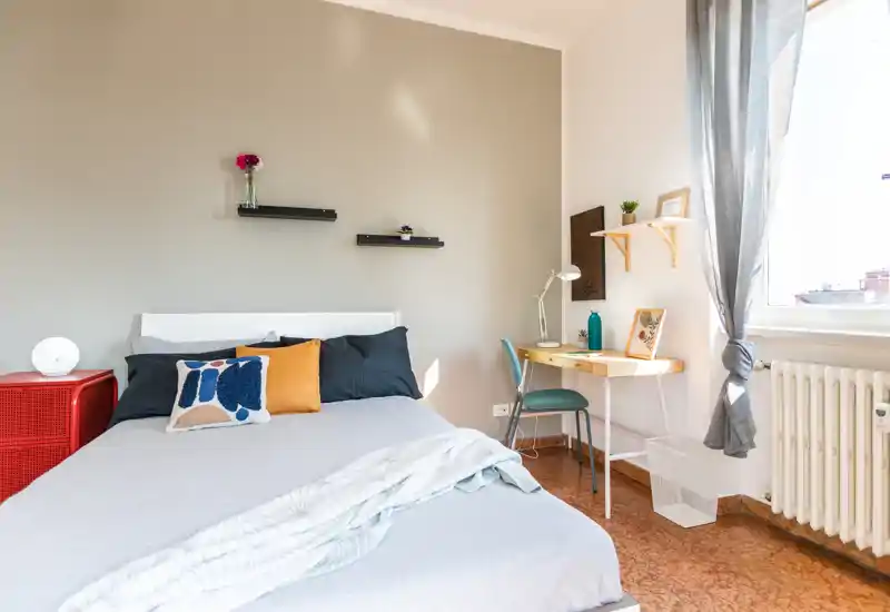 Camera da letto luminosa e ben arredata con un letto matrimoniale, cuscini decorativi e una piccola scrivania vicino alla finestra — ideale per un soggiorno confortevole con luce naturale.