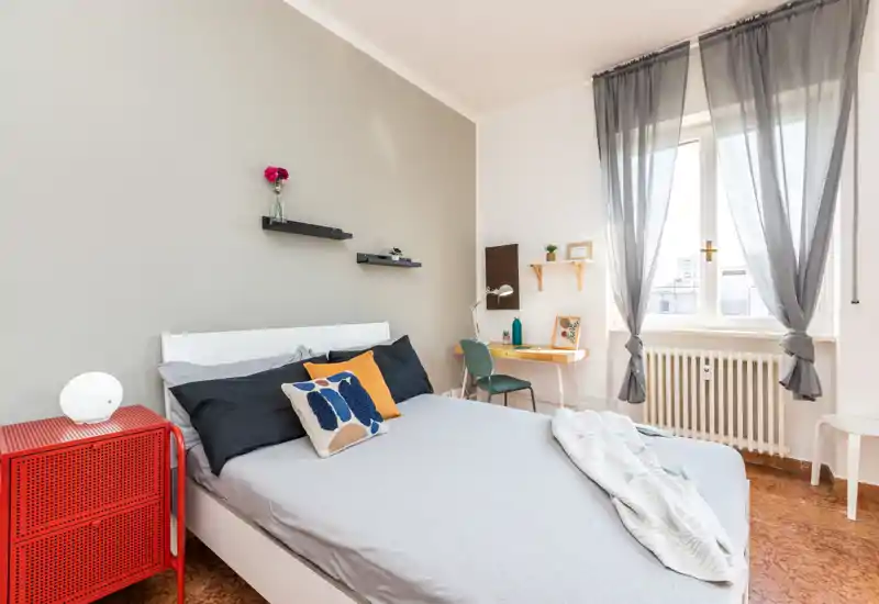 Camera da letto luminosa e ben arredata con letto matrimoniale, cuscini decorativi e un morbido plaid; ampia finestra con luce naturale e una piccola scrivania ideale per riposo e lavoro leggero.