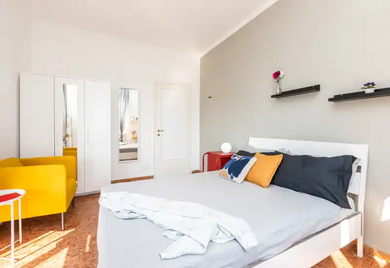 Chambre moderne et lumineuse avec un lit double bien fait, coussins colorés et un fauteuil jaune près de la fenêtre; étagères minimalistes et armoire pour un espace propre et accueillant.