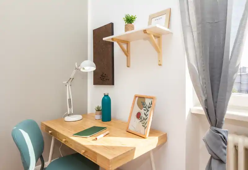 Coin bureau lumineux et soigné avec un bureau en bois, une lampe, un cadre décoratif et une petite plante près d'une fenêtre aux rideaux légers.