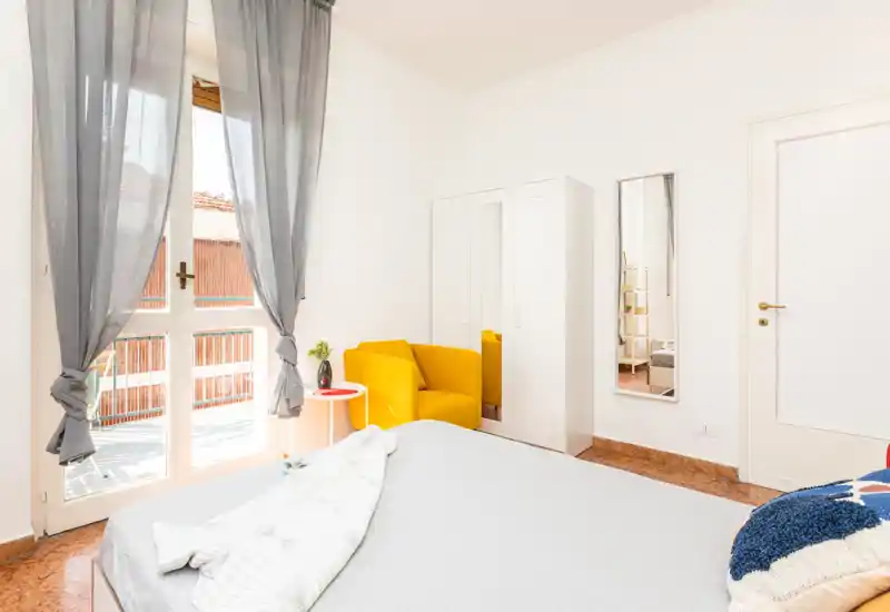 Camera da letto luminosa e curata con un ampio letto in primo piano, una poltrona gialla accanto all'armadio e porte-finestre che garantiscono molta luce naturale.