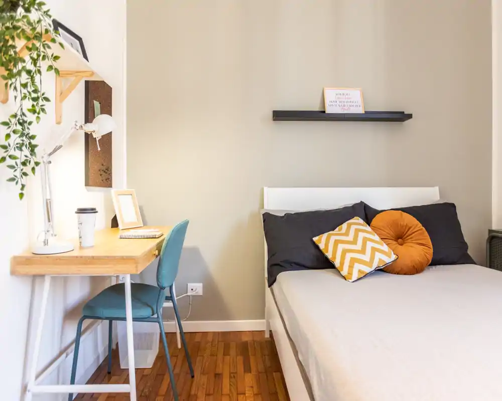 Camera da letto luminosa e ben arredata con letto matrimoniale in lino neutro e cuscini colorati; angolo studio compatto con scrivania in legno e sedia vicino alla finestra.