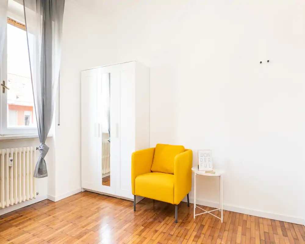 Angolo di camera luminoso e minimale con poltrona gialla, guardaroba con specchio e pavimento in legno — spazio semplice e arioso con luce naturale.