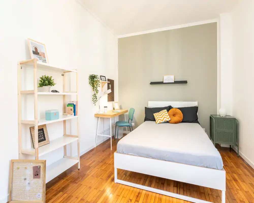 Camera da letto luminosa e curata con letto matrimoniale, cuscini decorativi, una piccola scrivania e scaffalatura — ideale per una persona sola o una coppia. La luce naturale e il pavimento in legno caldo creano un ambiente accogliente.
