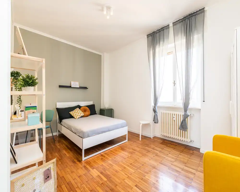 Camera da letto moderna e luminosa con letto matrimoniale ordinato, cuscini decorativi, pavimento in parquet e ampia finestra che garantisce molta luce naturale. Mensole semplici e un piccolo comodino completano lo spazio accogliente e ben arredato.