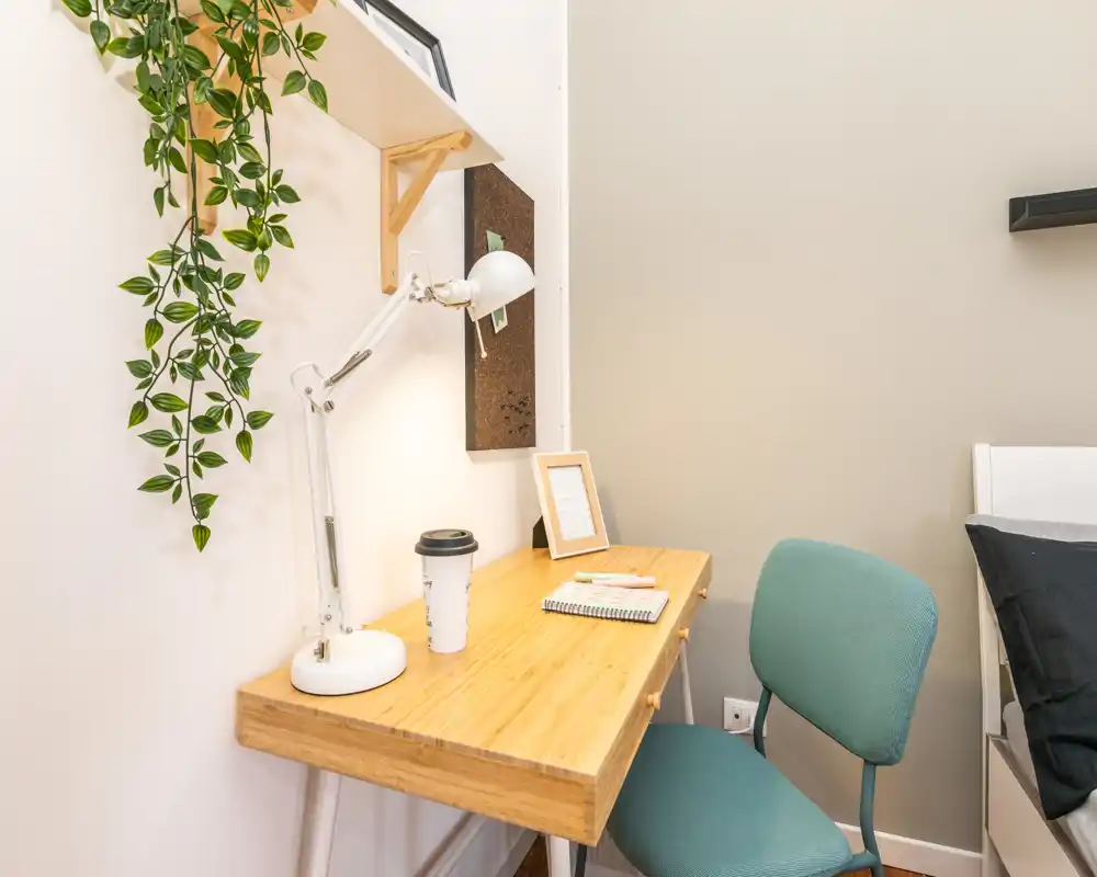 Angolo camera luminoso e ordinato con una scrivania in legno, lampada regolabile, quaderno e sedia accanto al letto — perfetto come postazione compatta.
