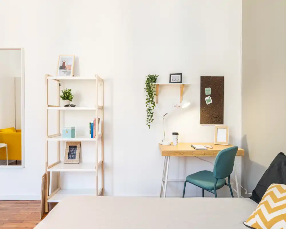 Angolo camera luminoso e moderno con letto in primo piano, una scrivania in legno con lampada e sedia turchese, scaffalatura aperta e piante decorative — ideale anche come piccolo spazio lavoro.