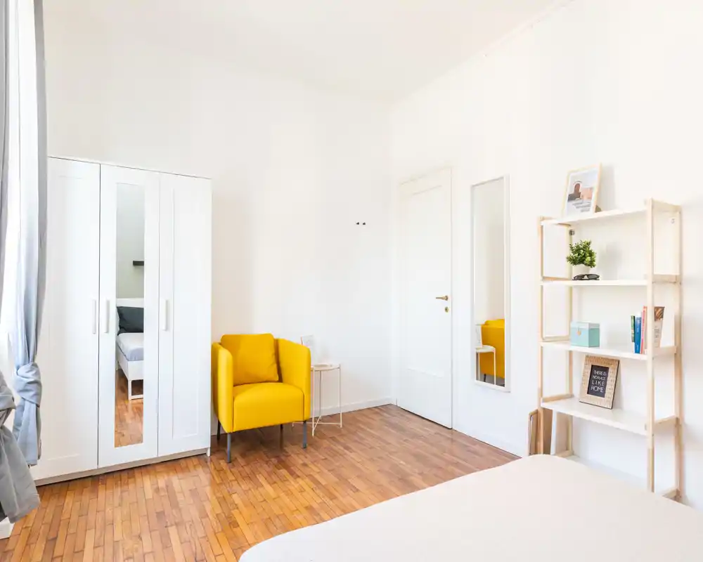 Camera da letto luminosa e minimal con vista parziale del letto, una poltrona gialla vivace, armadio bianco con anta specchiata e scaffalatura aperta — spazio arioso e pulito, ideale per una persona.