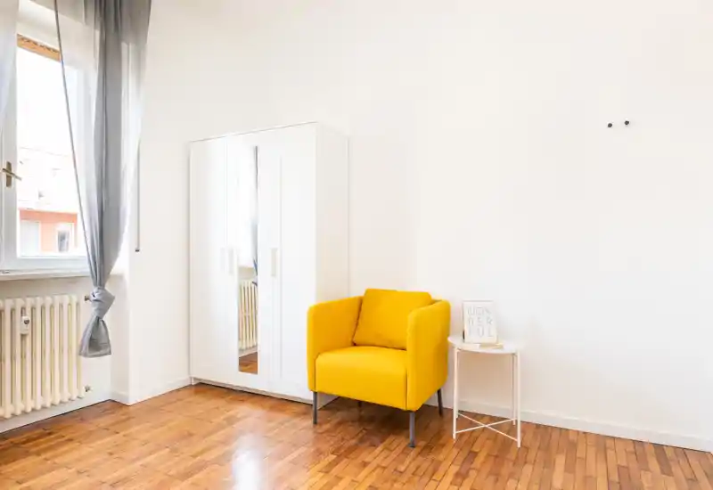 Coin de chambre lumineux et minimaliste avec un fauteuil jaune, une armoire miroir et un sol en bois — espace simple et aéré, lumineux naturellement.