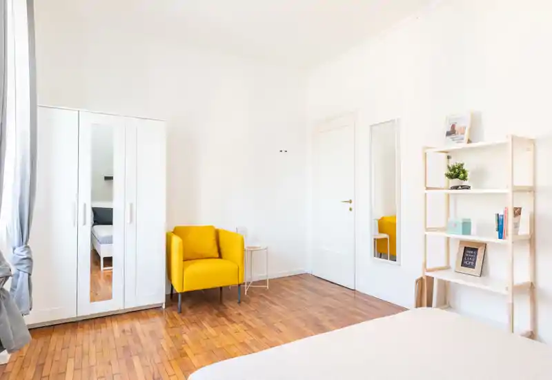 Chambre lumineuse et minimaliste avec vue partielle sur le lit, un fauteuil jaune vif, une armoire blanche avec porte miroir et des étagères ouvertes — espace propre et aéré, idéal pour une personne.