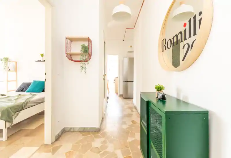Couloir lumineux et moderne avec une chambre ouverte sur la gauche ; la chambre présente un lit bien fait avec textiles verts et coussins, tandis que le couloir comporte un meuble vert et un miroir rond.
