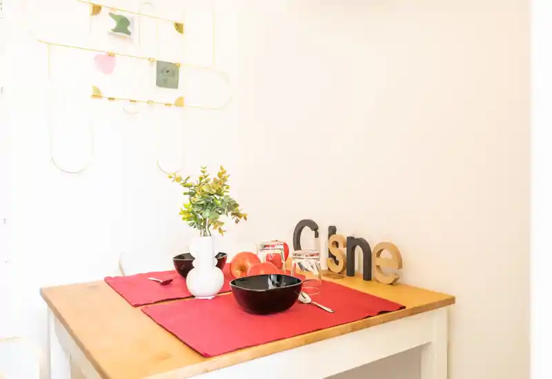 Coin repas intime avec table en bois dressée pour deux : sets de table rouges, bols noirs, vase décoratif avec verdure et décoration murale sobre. Endroit lumineux et chaleureux, parfait pour le petit-déjeuner.