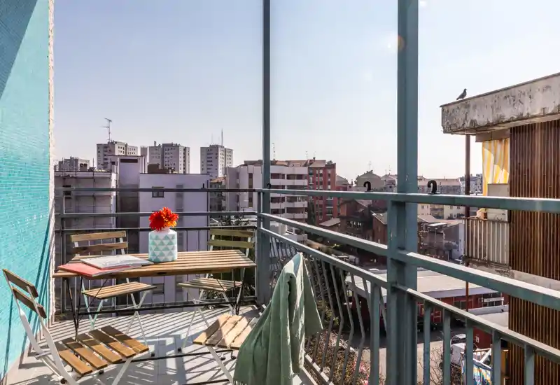 Balcon urbain ensoleillé avec petite table en bois et chaises pliantes, vase décoratif avec des fleurs et vue sur la ville — parfait pour le café du matin.
