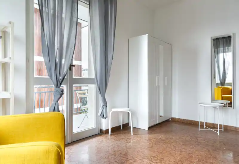 Zona de estar luminosa y moderna con luz natural, un sillón amarillo, armario y acceso a un pequeño balcón — aireada y amueblada con sencillez, ideal para un estudio.