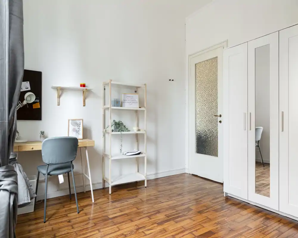 Rincón de dormitorio luminoso y minimalista con suelo de madera, armario blanco, escritorio y estantería: espacio ordenado para una sola persona.