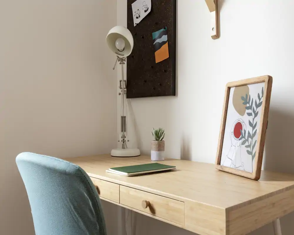 Un rincón de trabajo en casa ordenado con escritorio de madera, lámpara de mesa, cuadro enmarcado y una cómoda silla tapizada — ambiente luminoso y tranquilo, ideal para estudiar o trabajar desde casa.