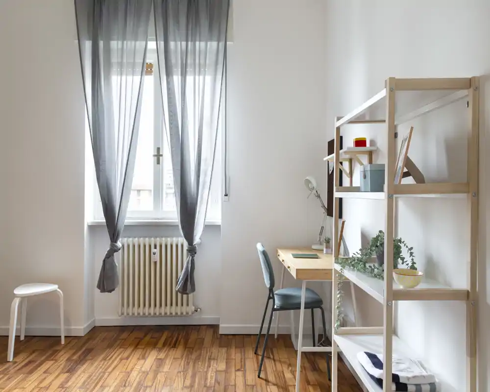 Zona de estar luminosa y minimalista con ventana, escritorio estrecho, estantería y planta acogedora — ideal para trabajar o relajarse en un apartamento compacto.
