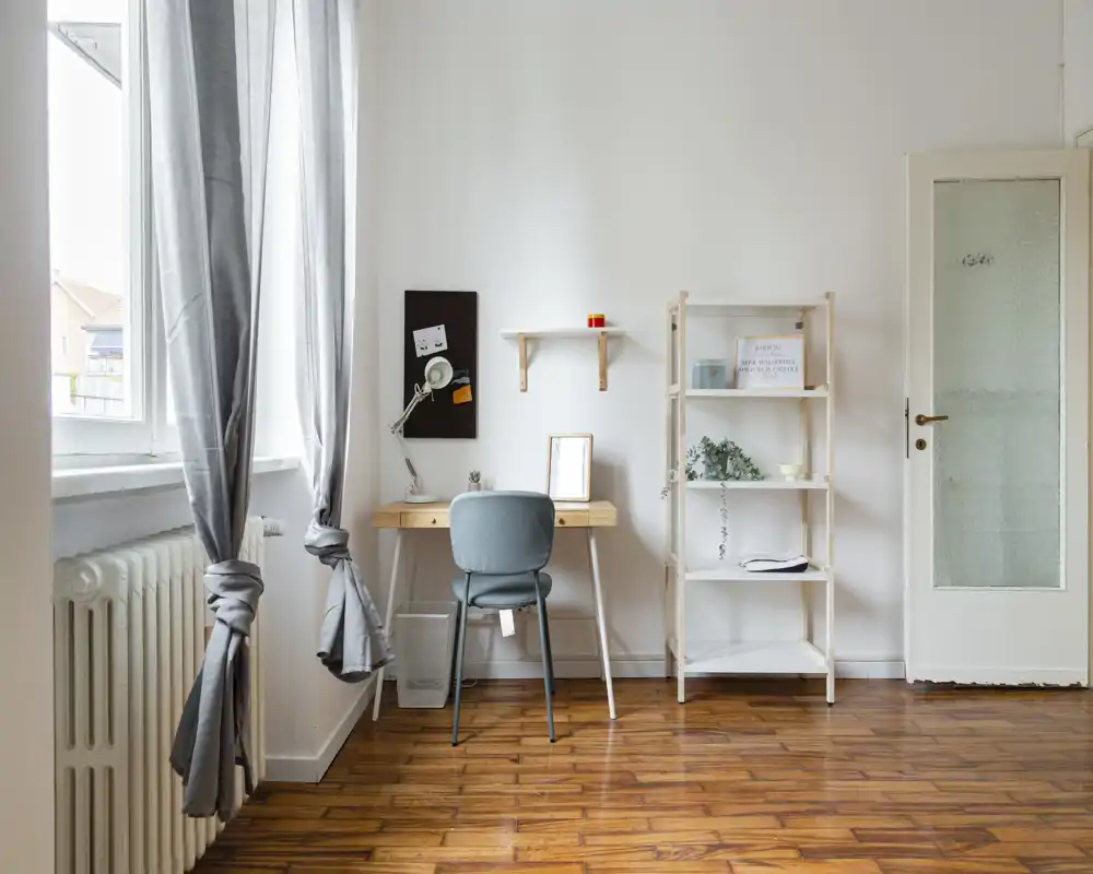 Zona de estar luminosa y minimalista con un pequeño escritorio y estantería junto a una gran ventana. La luz natural y la decoración sencilla crean un espacio tranquilo y funcional, ideal para trabajar o leer.