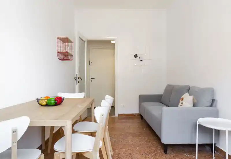 Zona de estar luminosa y compacta con sofá gris claro y mesa de madera con sillas blancas; estilo moderno y decoración minimalista, ideal para anunciar un piso pequeño.