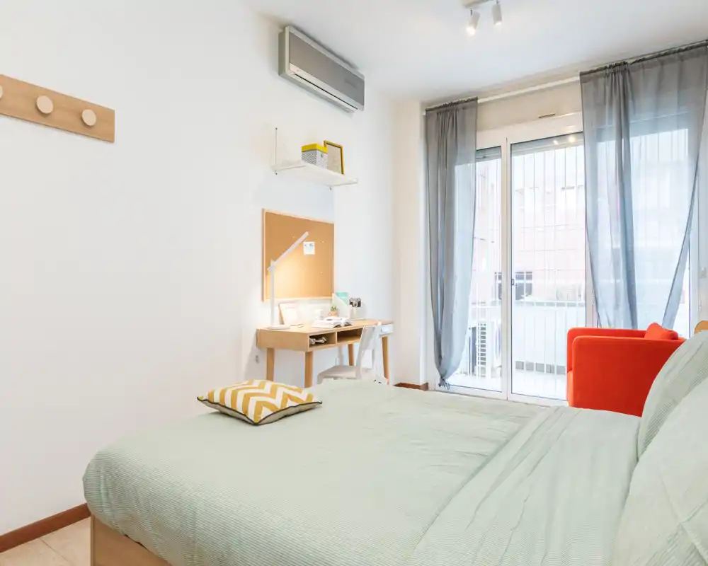 Dormitorio luminoso y bien dispuesto con una cama grande en primer plano, un pequeño escritorio de madera y acceso a un balcón por puertas correderas de cristal.