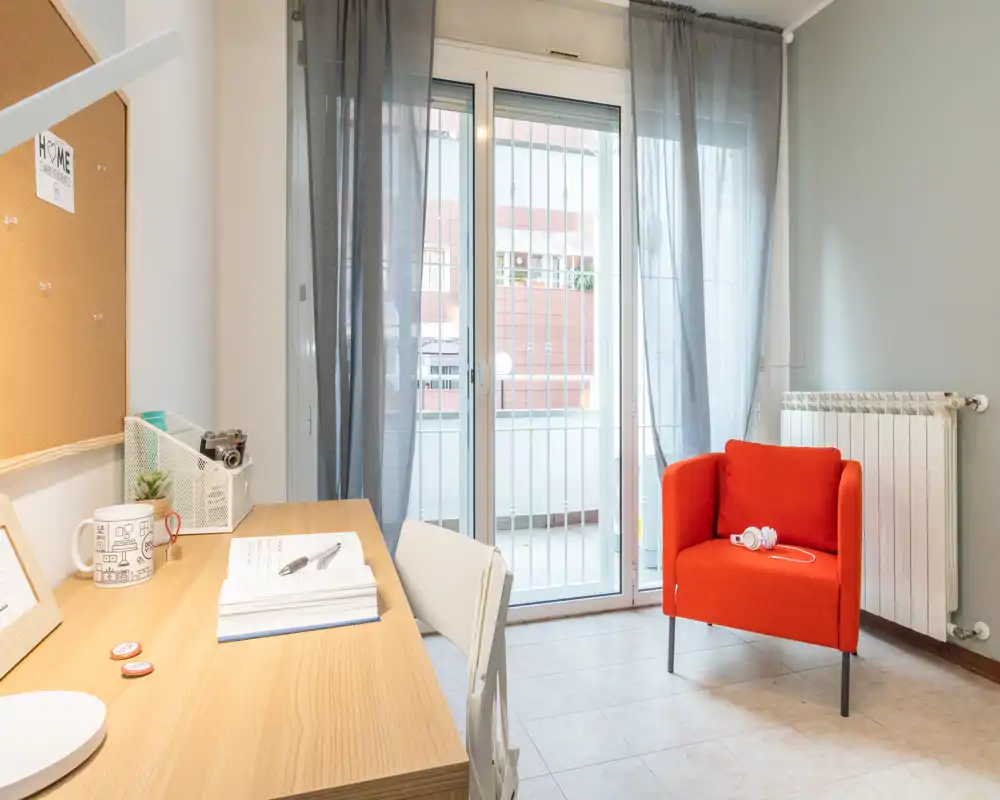 Rincón de estar moderno y luminoso con escritorio de madera, cómodo sillón rojo y grandes puertas acristaladas al balcón.