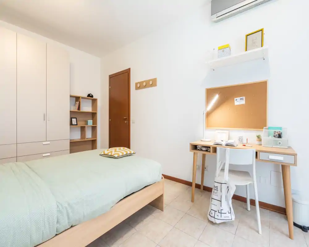Dormitorio individual luminoso y bien decorado con cama de madera, armario y escritorio. Tonos neutros, cama cómoda con cojín decorativo y un espacio de estudio práctico bajo un corcho.