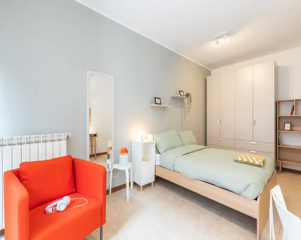 Dormitorio moderno y luminoso con cama doble vestida con ropa de cama verde suave, un gran armario y un sillón naranja que aporta un toque de color. Estanterías sencillas, espejo de pie y luz cálida en la mesita, ideal para anuncios de alquiler que destaquen confort y estilo.