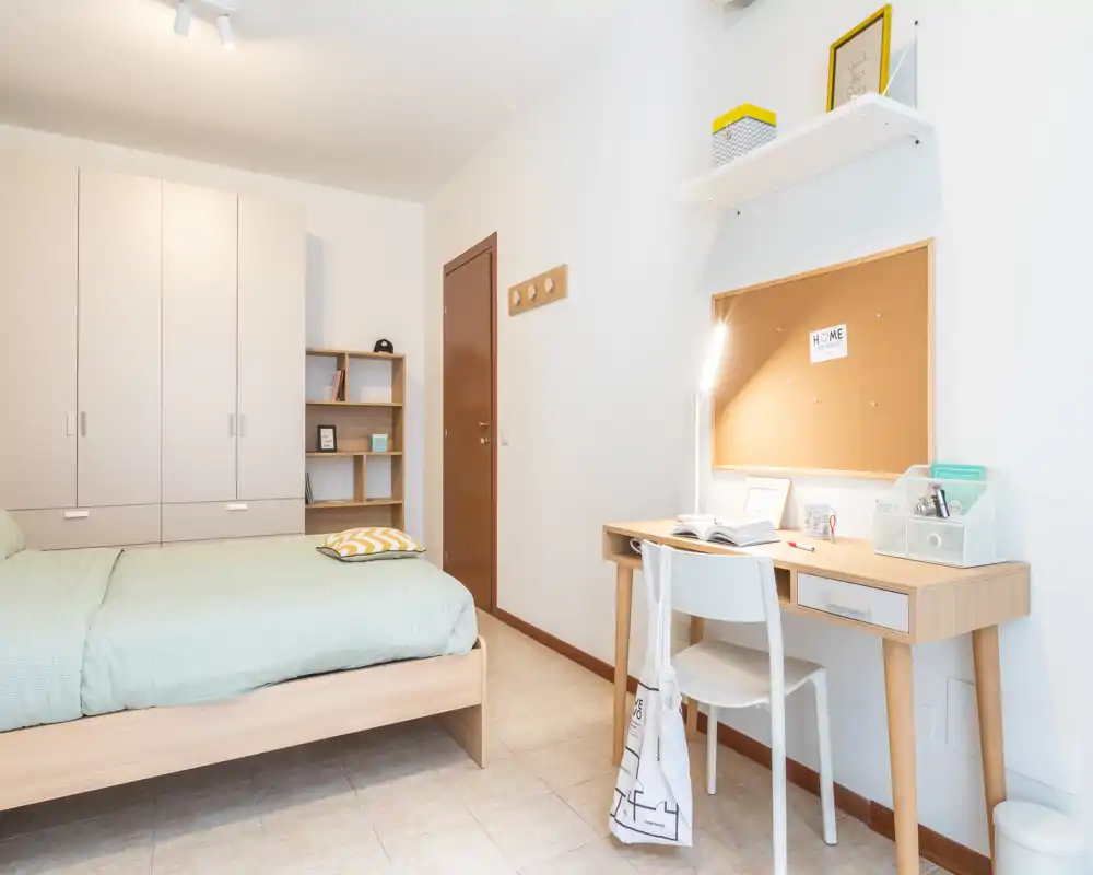 Dormitorio luminoso y minimalista con cama de madera, armario y un pequeño escritorio de estudio. El espacio es fresco y bien iluminado, con líneas limpias y textiles claros.