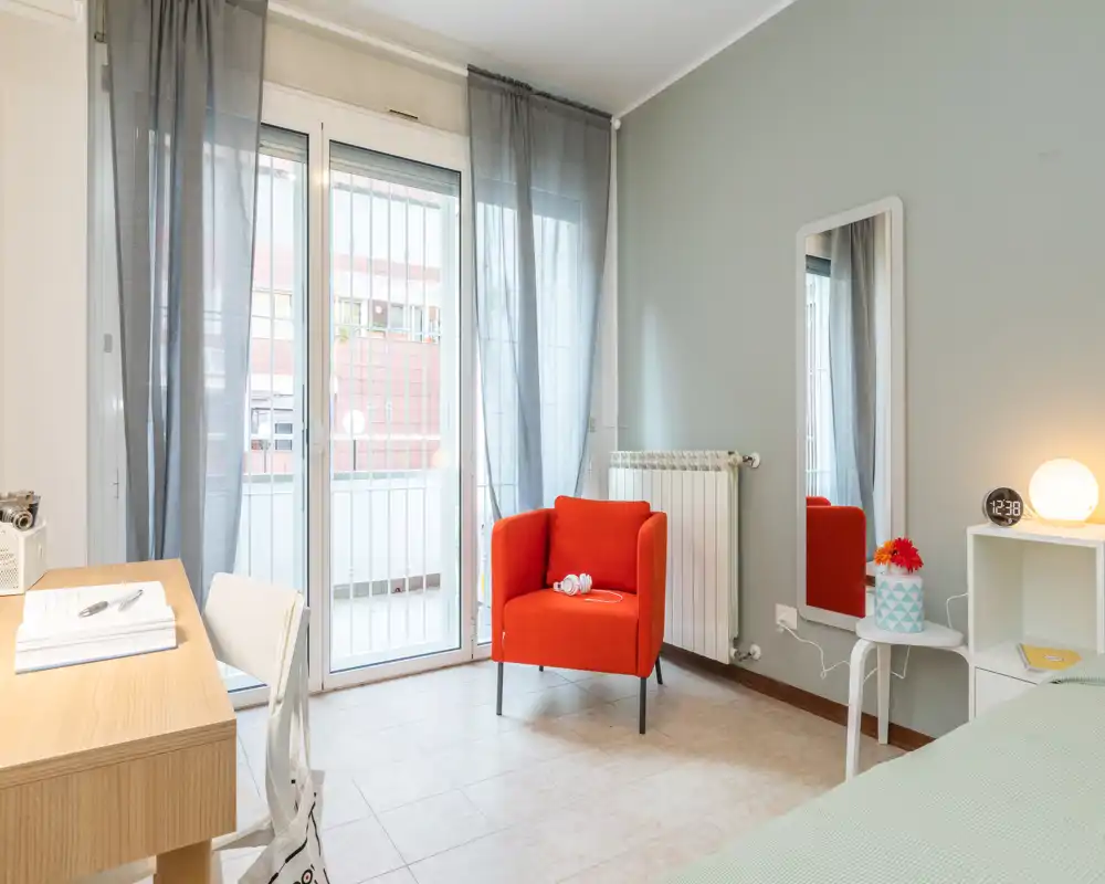 Rincón de dormitorio luminoso y moderno con un sillón rojo de acento, escritorio junto a la ventana y una porción de la cama visible. Grandes puertas al balcón aportan luz natural y cortinas translúcidas.