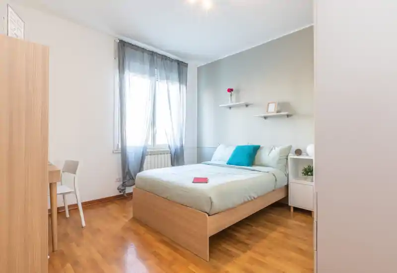 Dormitorio luminoso y moderno con cama doble, suelo de madera clara y toques en turquesa. Cortinas translúcidas, estantes sencillos y mesita ordenada ofrecen un ambiente acogedor y minimalista.